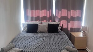 Una cama bien hecha con ropa de cama estampada, una mesita de noche con lámpara y cortinas que cubren las ventanas.