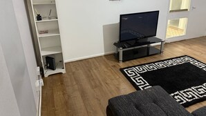 Una sala de estar con un televisor de pantalla plana, un estante blanco, un sofá negro y una alfombra estampada.