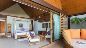 Een moderne hotelkamer met een grote schuifglazen deur die uitkomt op een balkon, een bed met hemelbed, een zithoek met een bank en stoelen, en een kleine tafel.