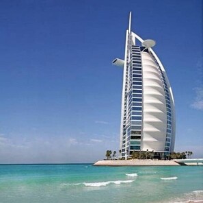A tall, white, curved building with a sail-like structure, located on a small island with clear blue water and a sandy beach.