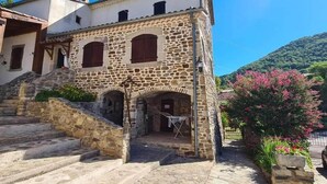 A stone building with a staircase leading to a door, surrounded by a cobblestone courtyard and a hillside in the background. - Mes Maisons Charming 3-bedroom Gite with AC, fitness room in delightful Cevennes (Gagnières)