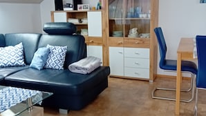 A modern living room with a sofa, a glass coffee table, a dining table with chairs, a cabinet with glass doors, and a wooden ceiling. - Holiday Apartment 60sqm, 2 Bedrooms, Max. 4 Persons, 4 Stars - DTV Classified (Hayingen)
