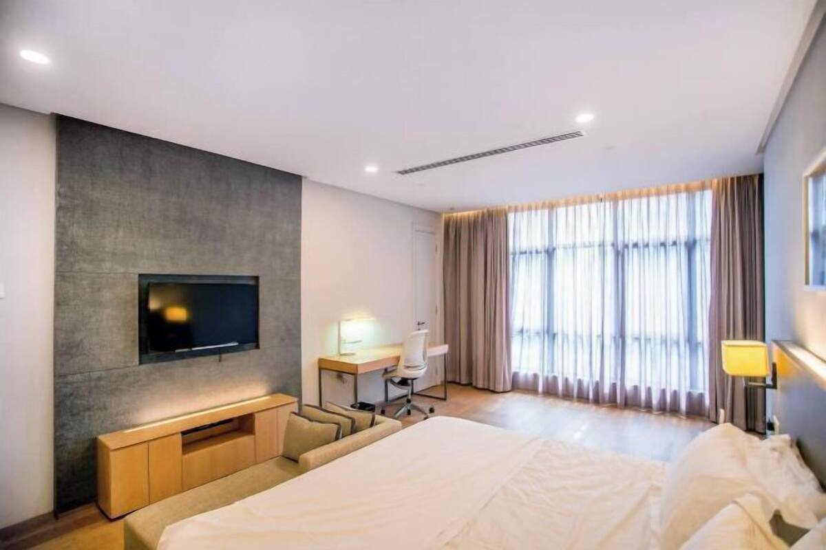 Een moderne hotelkamer met een bed, een bureau, een aan de muur bevestigde televisie en grote ramen met gordijnen.