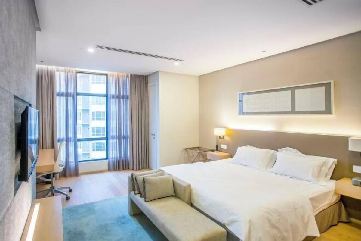 Hotelkamer met een bed, bureau, stoel, bankje, televisie en raam met gordijnen.