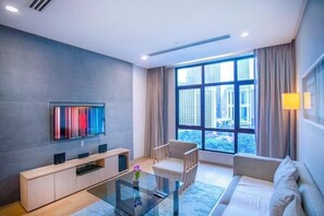 Een moderne woonkamer met een bank, een glazen salontafel, een aan de muur bevestigde televisie en een groot raam met gordijnen.