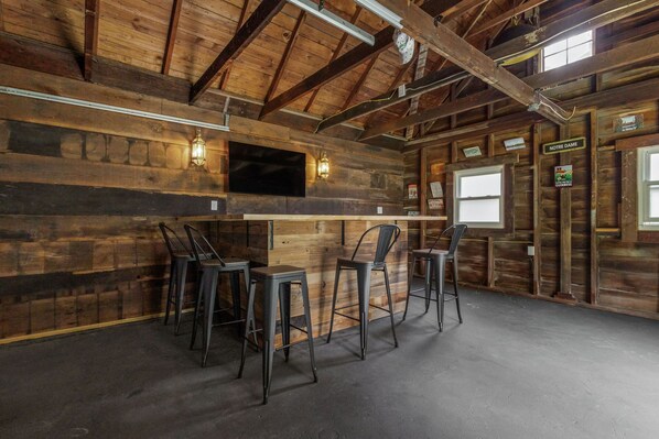 Een rustiek interieur met een bar, metalen krukken en een flatscreen televisie bevestigd aan een houten wand.