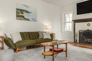 Een woonkamer met een groene bank, een ronde houten tafel, een open haard en een schilderij aan de muur.