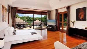 A spacious bedroom with a large bed, a television, and a view of a garden and building through a sliding glass door. - Laguna Waters - Lake View Villa, 5 Bedrooms (Bang Tao)