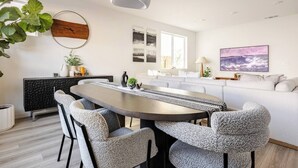 A modern dining area with a round table, grey upholstered chairs, a black console, and a white pendant light.
