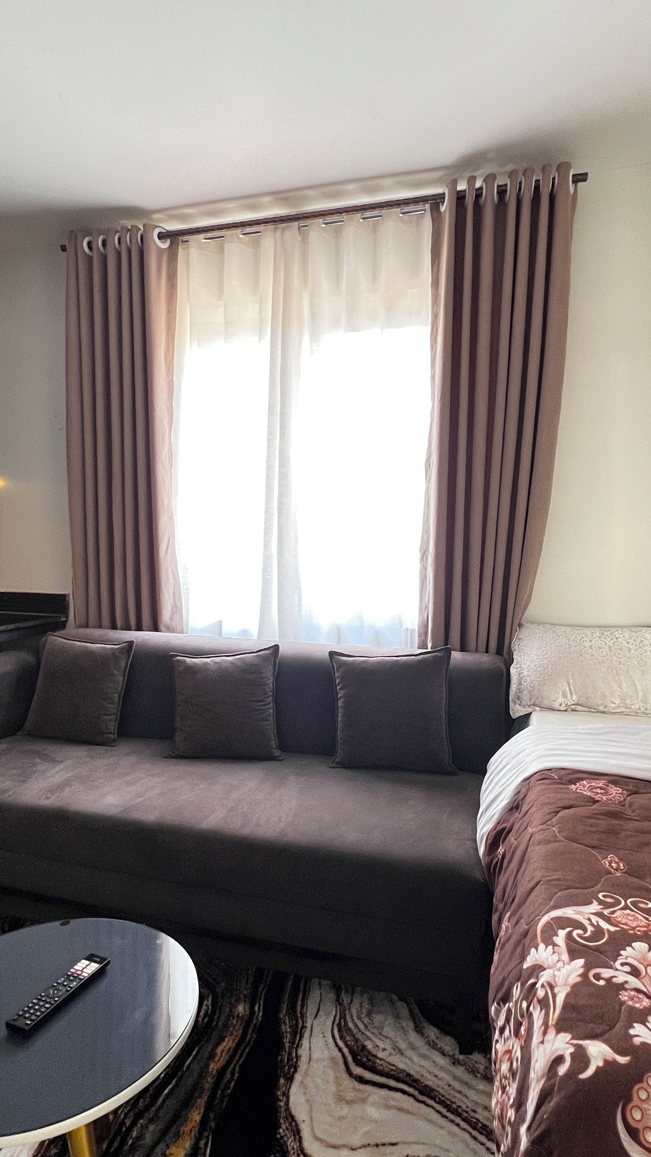 A hotel room with a grey sofa, a bed with patterned bedding, a round coffee table with a remote control, and a window with curtains.