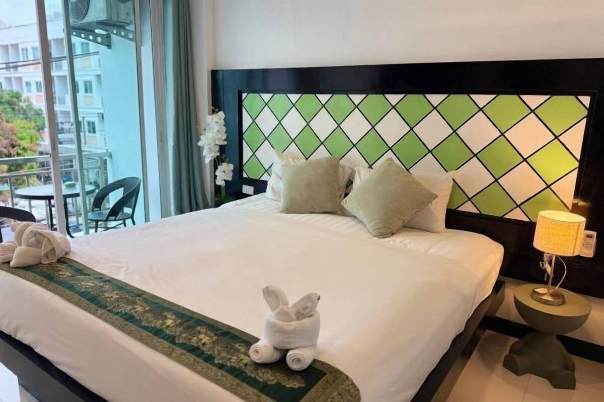A hotel room with a bed, a headboard with a geometric pattern, a nightstand with a lamp, and a view of the outside balcony.