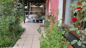 Área de varanda coberta com mesa e cadeiras, cercada por vegetação e uma parede vermelha.
