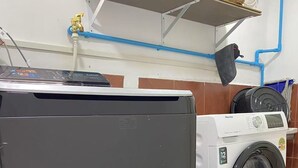 Una lavadora y secadora compactas con panel de control, ubicadas sobre un piso de madera con tuberías visibles y un ventilador de techo al fondo.