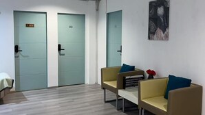 Un área de espera moderna con tres puertas identificadas con los números 20, 03 y 02. Hay dos sillones amarillos con cojines azules y una pequeña mesa con un florero rojo.