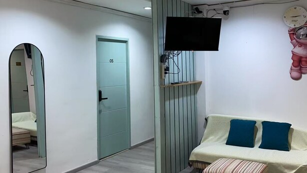 Una habitación moderna con un espejo grande, un televisor montado en la pared, un sofá con cojines y una puerta con la etiqueta '05'.