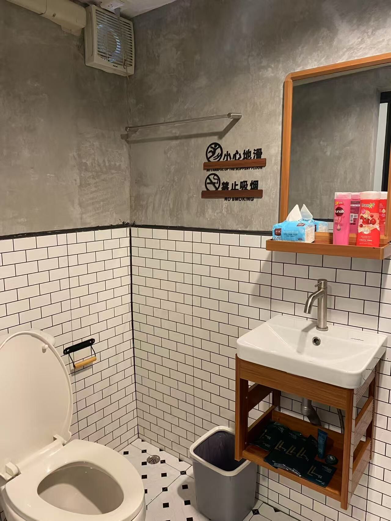 A small, tiled bathroom with a toilet, sink, and mirror. There is a trash can and a shelf with toiletries.