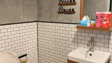 A small, tiled bathroom with a toilet, sink, and mirror. There is a trash can and a shelf with toiletries.