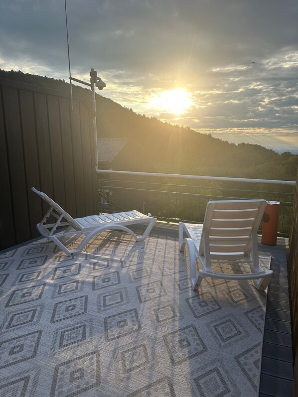 A balcony with two white chairs and a patterned rug, overlooking a hilly landscape with a sunset.