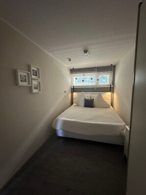A small, narrow room with a bed, a window with blinds, and three framed pictures on the wall.