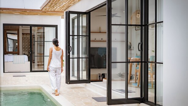 A modern indoor pool area with a clear glass wall, a wooden ceiling, and a visible bedroom with a bed and a window. - Villa Lumière 1 Seminyak (Seminyak)