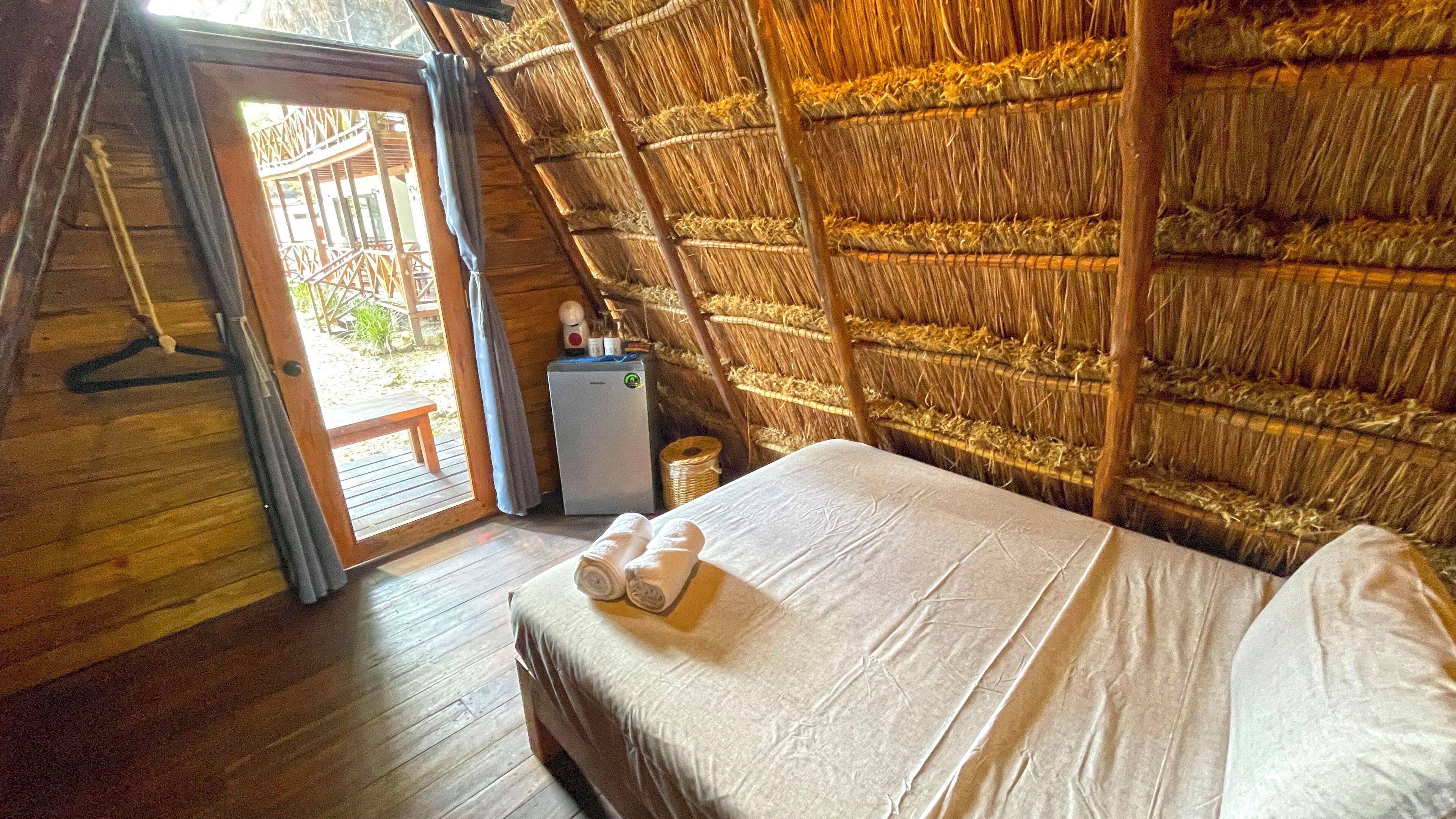 Quarto com piso de madeira, cama com roupa de cama branca, uma pequena mesa com uma garrafa e um recipiente, e um telhado de palha.