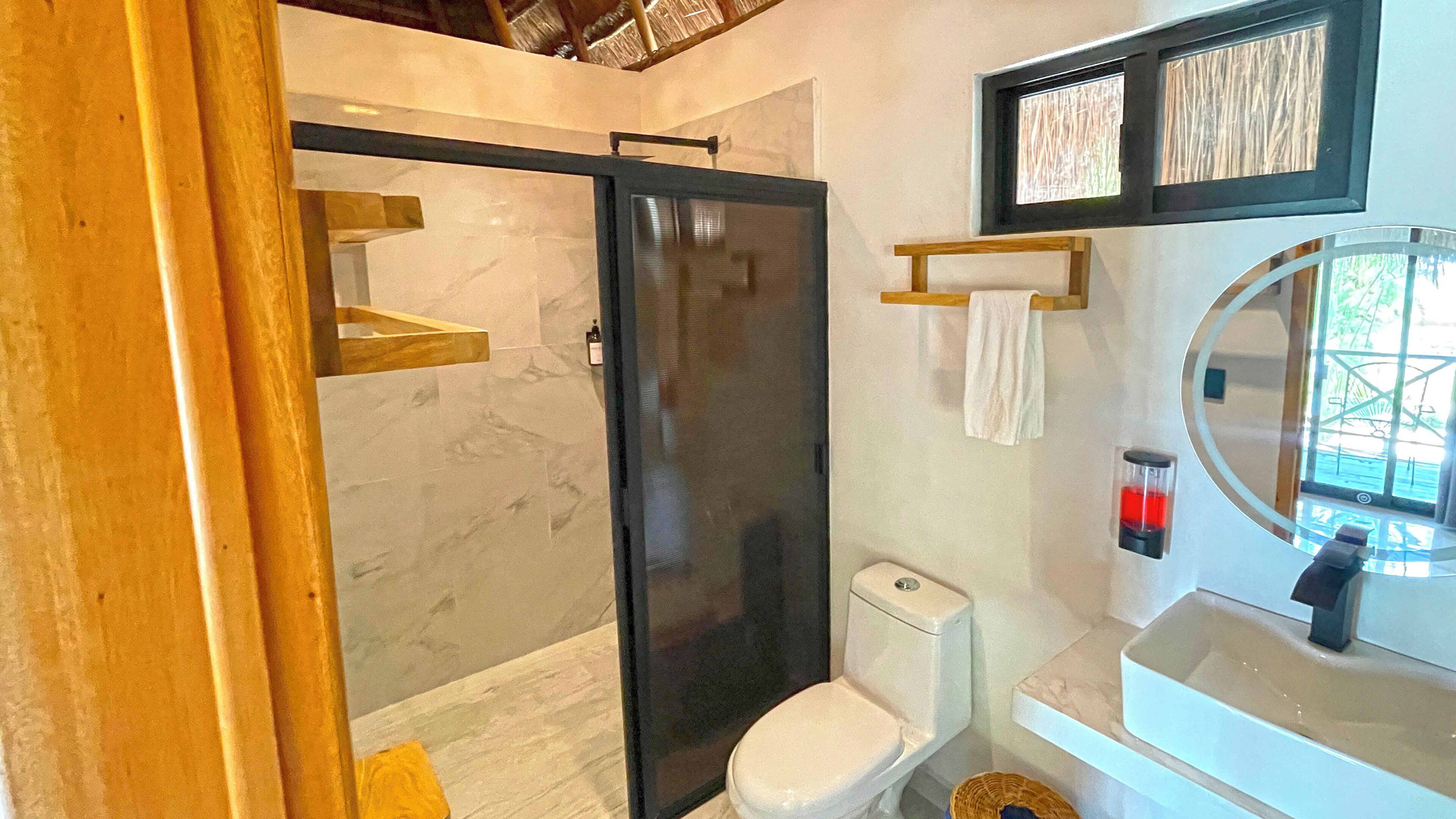 A modern bathroom with a glass shower enclosure, a toilet, a sink with a mirror, and a window with a view.