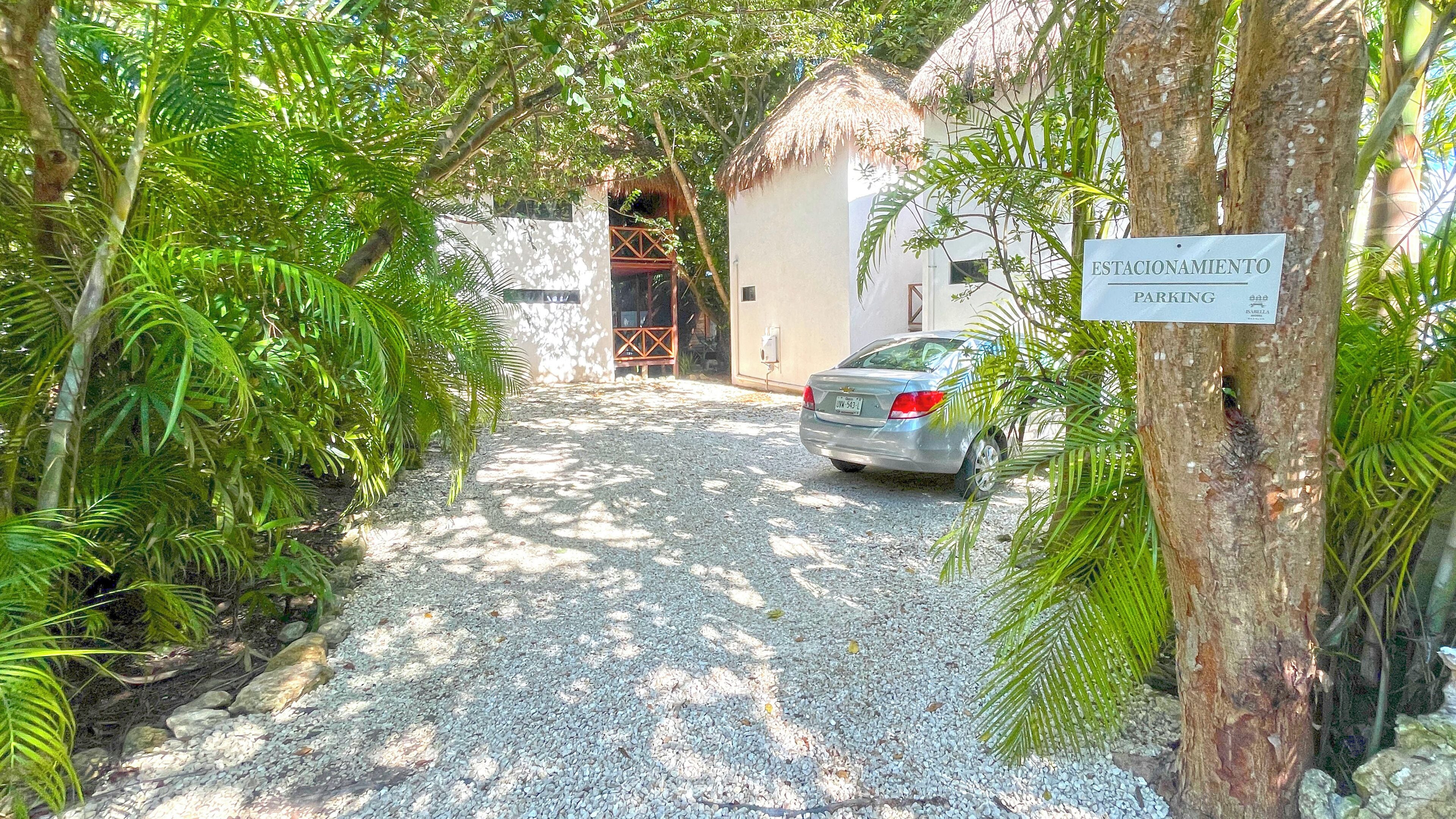 Uma entrada de cascalho leva a um edifício com telhado de palha, cercado por vegetação exuberante.