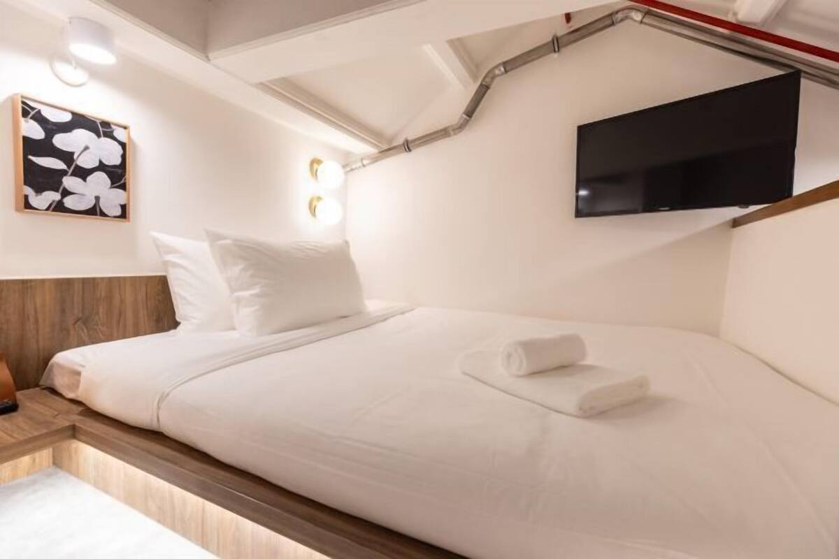 A single bed with white bedding, a mounted TV, a wall-mounted light fixture, and a floral artwork on the wall.