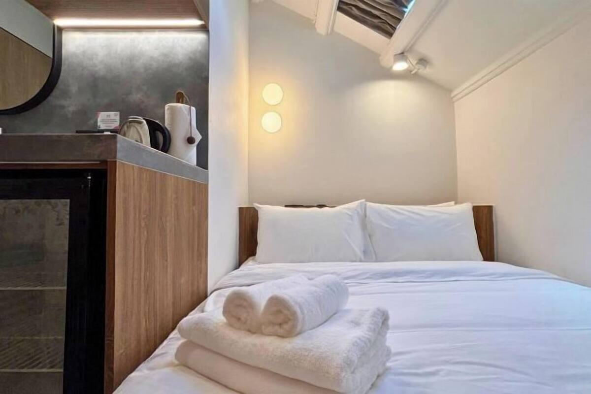 A hotel room with a bed, a wooden headboard, a nightstand with a kettle, and a wall-mounted light.