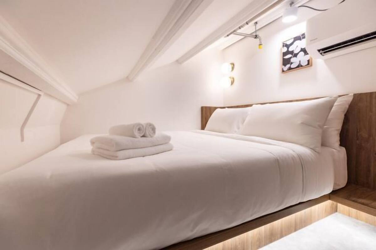 A neatly made bed with white linens and a folded towel, a wooden headboard, and a wall-mounted light fixture.