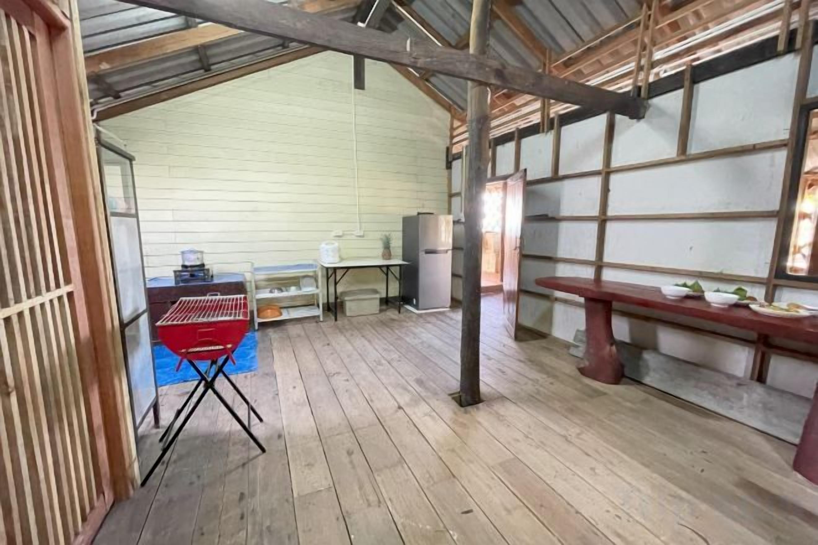 Un interior rústico con suelo de madera, una parrilla roja, un banco y una mesa con platos.