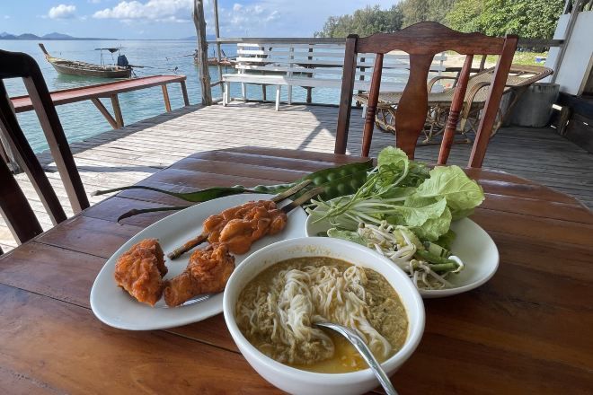 A table with a bowl of noodle soup, a plate of fried chicken, and a plate of fresh greens, set on a wooden deck overlooking a body of water.