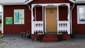 A small, single-story building with a wooden door labeled "ENTRE" and a porch with white railings. There are two flower pots on the steps and a map on the wall to the left. - 4 Person Holiday Park Home in 82011 (Vallsta)