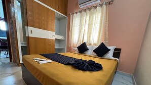 A neatly made bed with a yellow bedspread and two black pillows, a wooden wardrobe, and a window with sheer curtains. - Surya-Atharva Suites (Mysore)