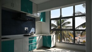 A modern kitchen with teal cabinets, a white countertop, and a window with a view of palm trees. - Surya-Atharva Suites (Mysore)