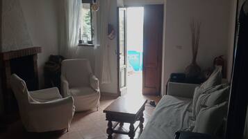 A living room with a fireplace, two white armchairs, a wooden coffee table, and a sofa. There is a door leading outside.