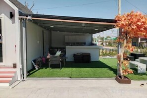 Un espacio al aire libre cubierto con un pequeño césped artificial, una zona de estar y un árbol de hojas anaranjadas.