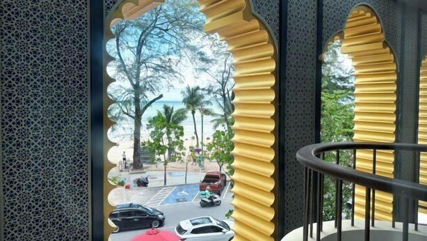 Vista a través de una ventana con un marco decorativo, barandilla de balcón y una calle con coches aparcados y palmeras.