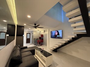 Un salon moderne avec un canapé en cuir noir, une table basse blanche et un téléviseur fixé au mur. Un escalier avec une rampe noire mène à l’étage supérieur.