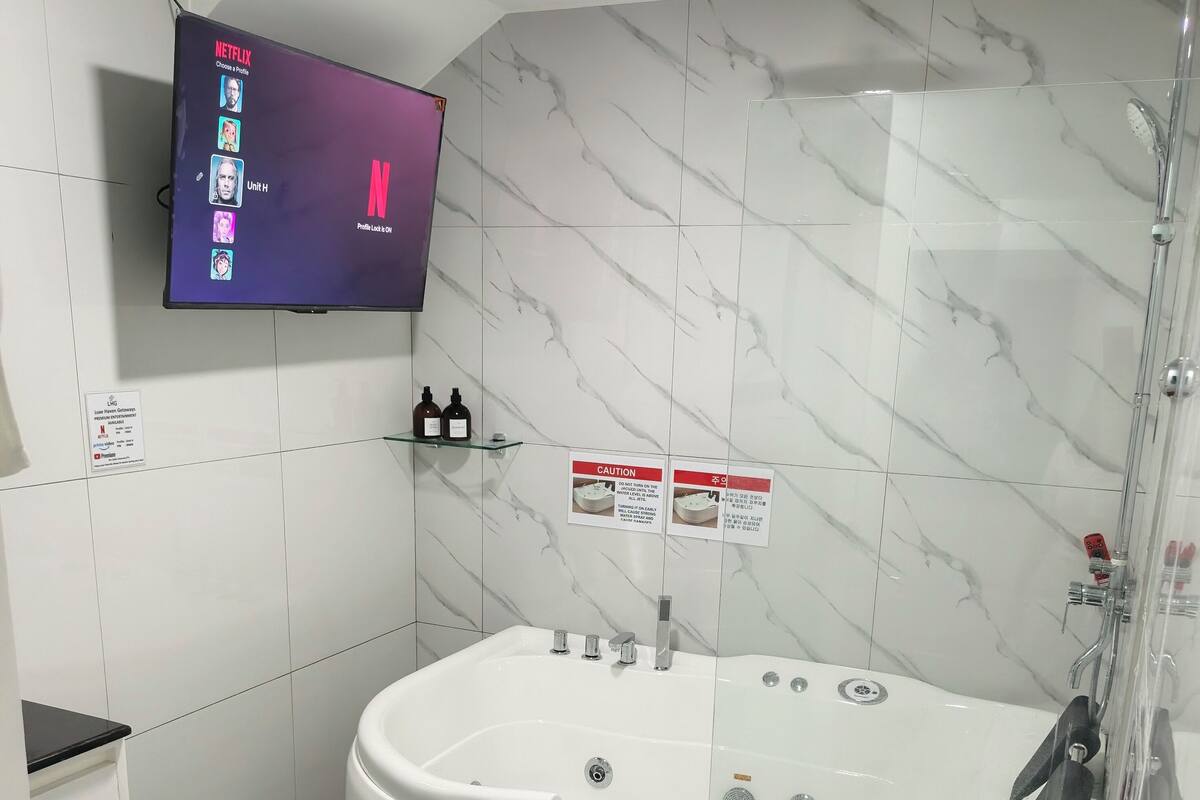 A modern bathroom with a large bathtub, a toilet, a shower, and a wall-mounted TV displaying Netflix profiles.