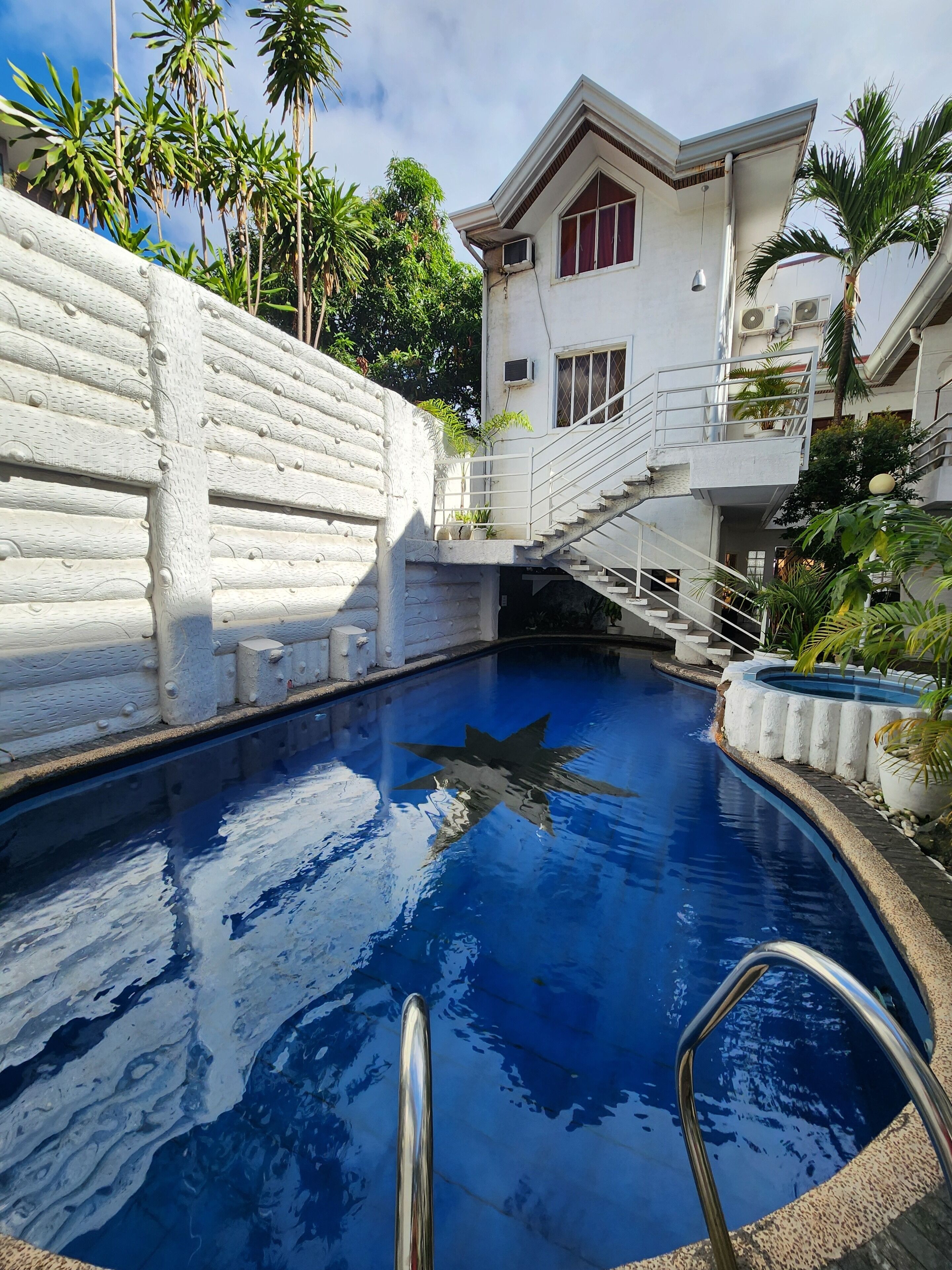 A swimming pool with a white stone wall, a staircase, and a building with a red window.