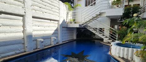 A swimming pool with a white stone wall, a staircase, and a building with a red window.
