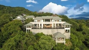 A large, multi-story house with a flat roof, surrounded by dense greenery and hills in the background.