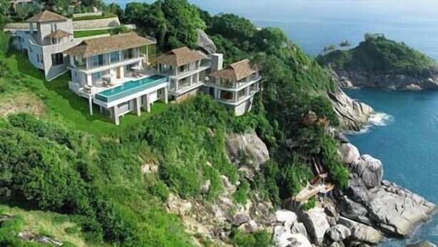 A large, modern house with multiple levels and a pool, surrounded by lush greenery and overlooking a coastal area.