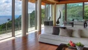 A spacious living room with a large white sofa, a coffee table, and a view of the ocean through floor-to-ceiling windows.