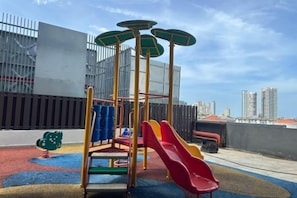A playground with a slide and climbing structure, surrounded by a fenced area with a cityscape in the background. - JazzA Suite by Homey Planet (Tanjung Tokong)