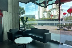 A modern lobby with a black sofa, a round table, and a large window offering a view of a building and a street. - JazzA Suite by Homey Planet (Tanjung Tokong)