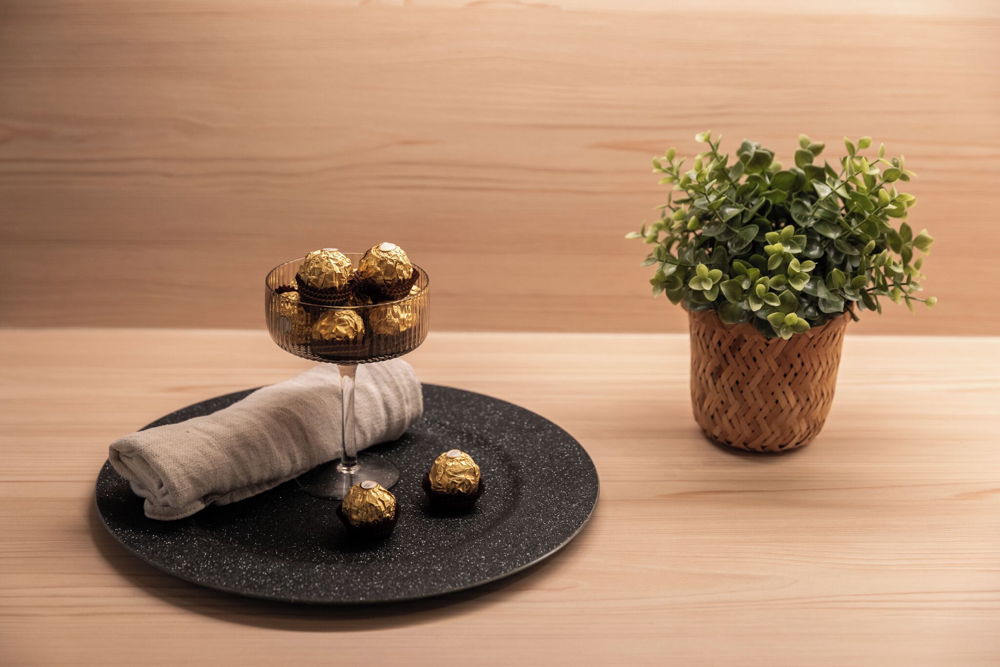 Une assiette noire ronde, sur laquelle repose une serviette en tissu pliée, un bol en verre contenant des chocolats emballés dans du papier doré, et une petite plante en pot, le tout posé sur une surface en bois.