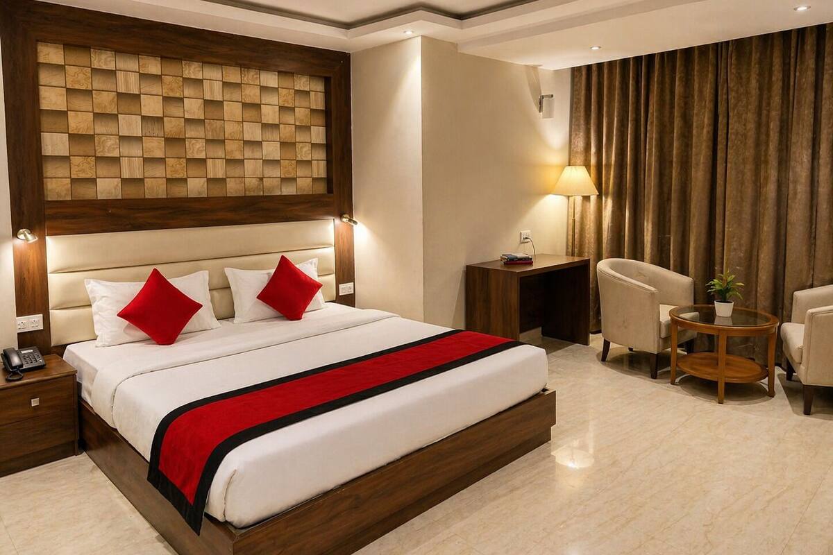 Habitación de hotel con una cama grande, cabecera de madera, mesita de noche con lámpara, una silla y una pequeña mesita con una planta.