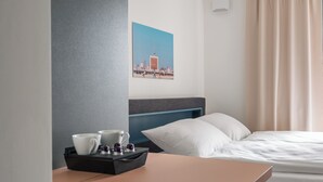 Ein Hotelzimmer mit einem Bett, einem Nachttisch mit Tablett, Tassen und einer kleinen Flasche, einem Kissen und einem an der Wand hängenden Stadtbild.
